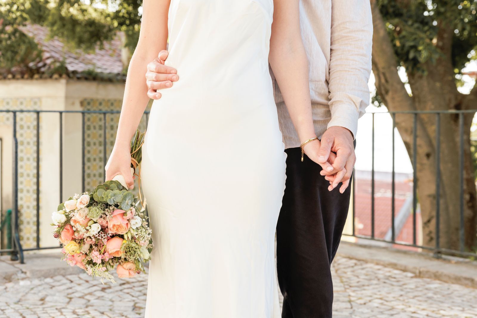 Bride and groom holding hands during their Lisbon elopement, with the bride holding a pastel floral bouquet and wearing a minimalist white dress in a sunlit historic setting.