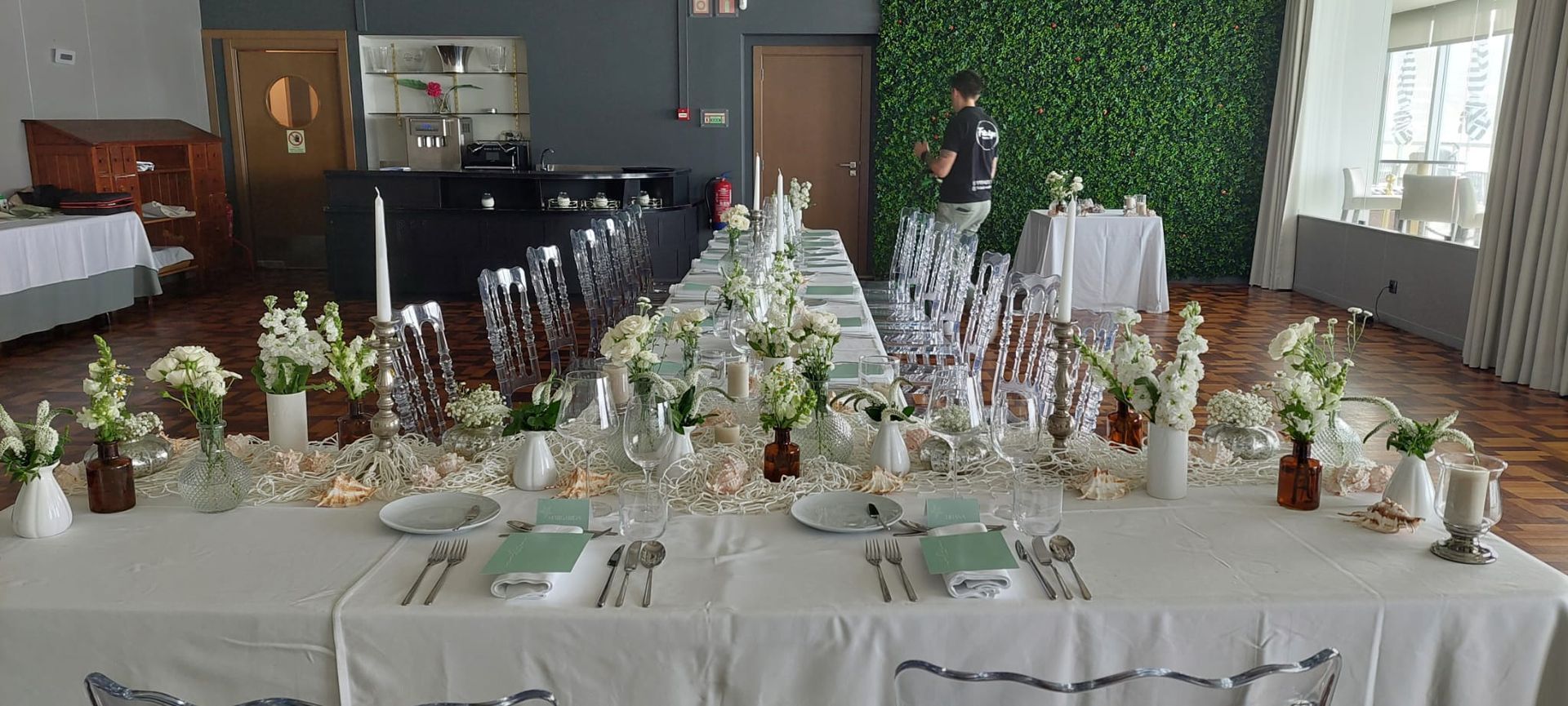A shot of a long, elegantly set reception table in a modern space with green and white decor, ready for a formal dinner. A staff member is seen making final preparations in the background. Hotel Arribas at Praia Grande, Colares.