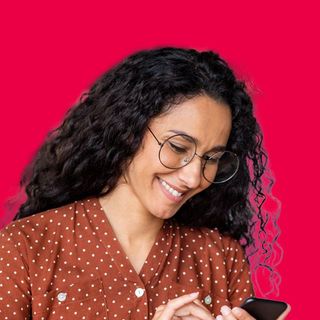 Smiling person with curly hair and glasses looking at a smartphone on a pink background
