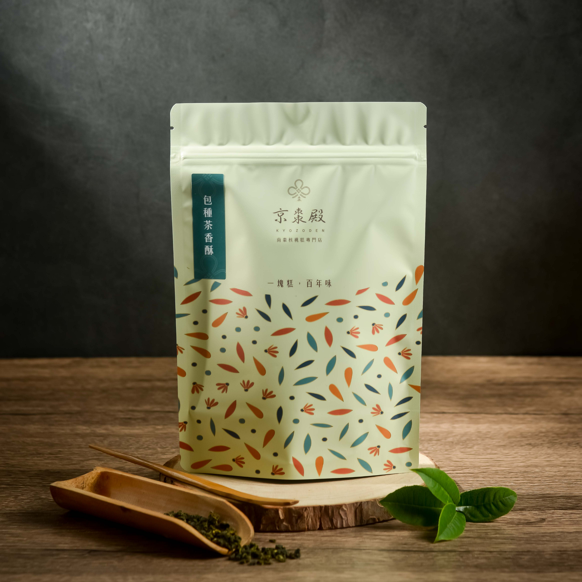 木質表面上放著一個米白色茶包，上面印有色彩繽紛的葉子圖案，旁邊還配有一個茶勺和一小枝薄荷葉。