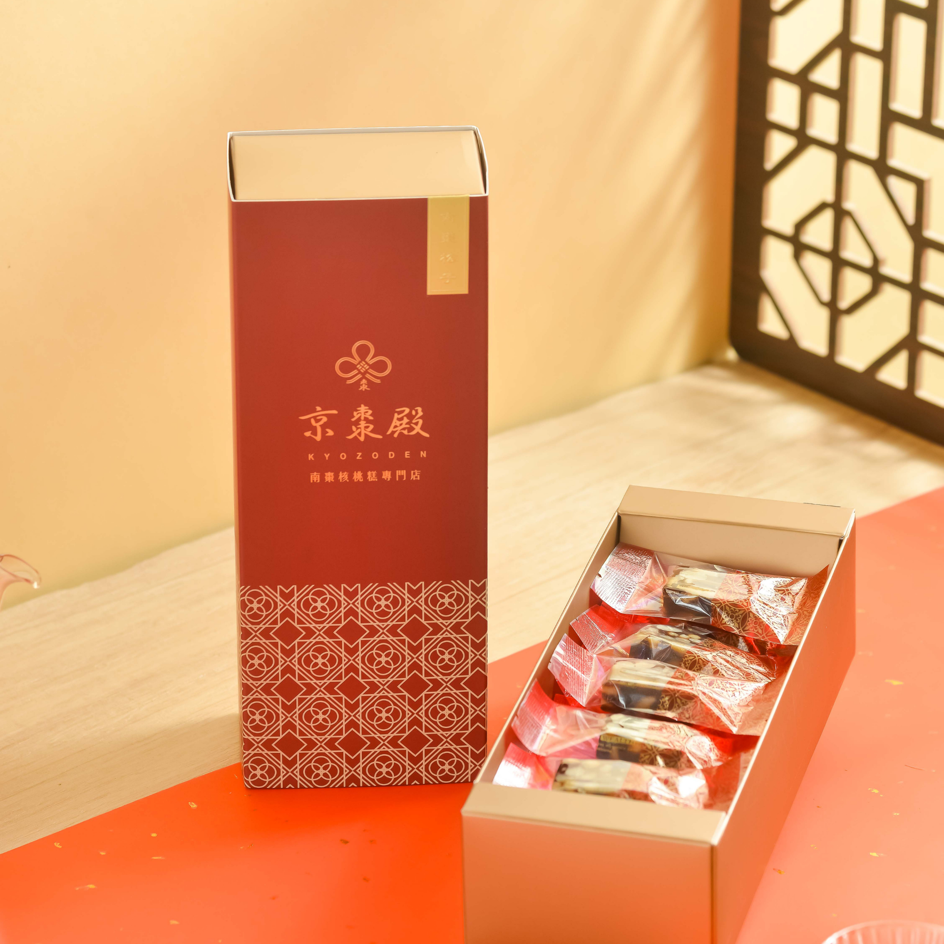 木托盤上放著玻璃茶壺和茶杯，旁邊放著一個紅色禮盒，周圍環繞著茶葉，背景較暗。