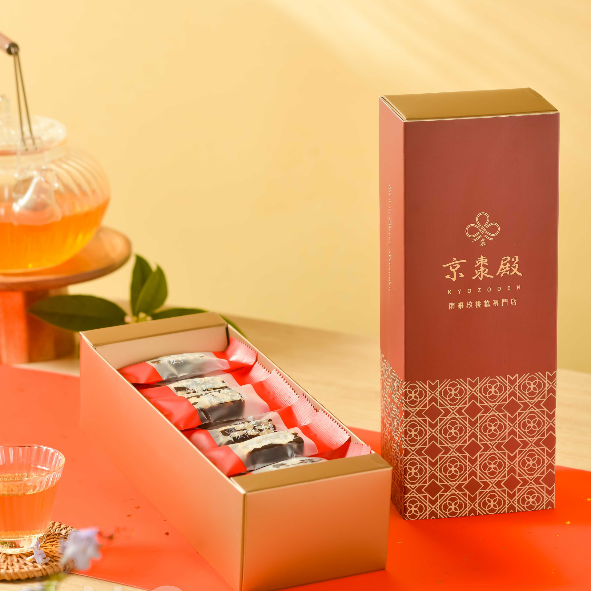 暖黃色背景上的紅色茶禮盒，置於木托盤上，內含幾小杯琥珀色茶和柑橘。