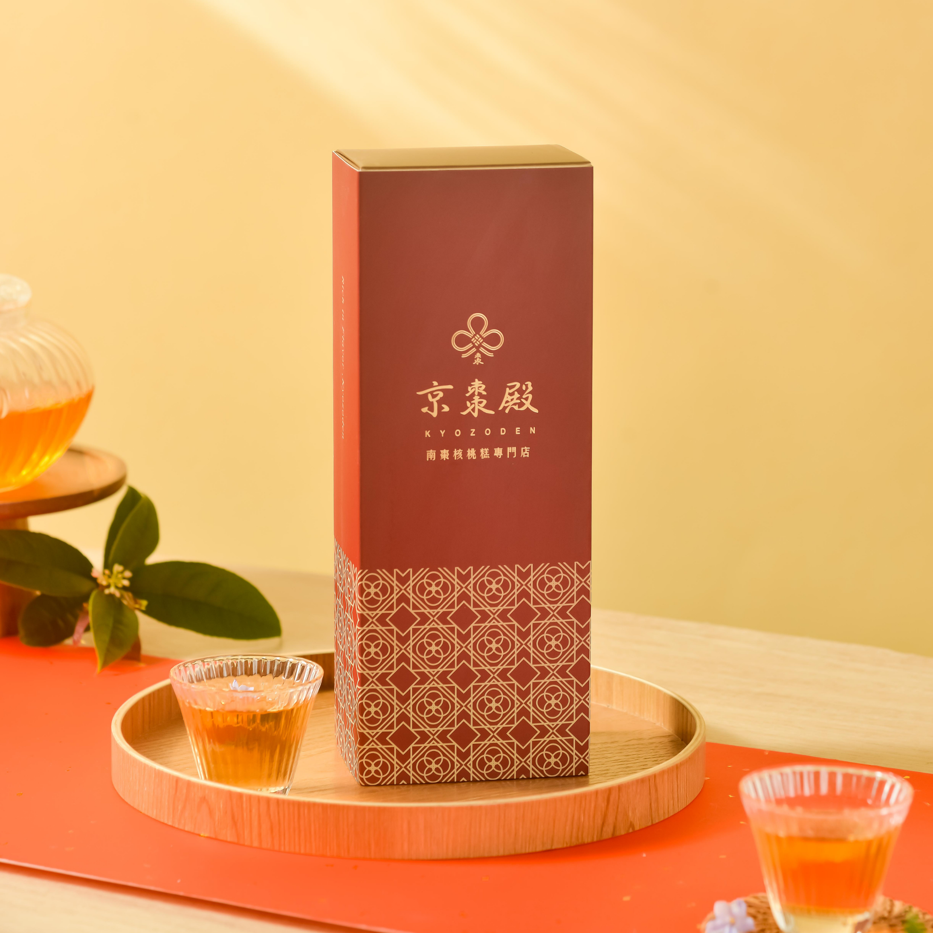 暖黃色背景上的紅色茶禮盒，置於木托盤上，內含幾小杯琥珀色茶和柑橘。