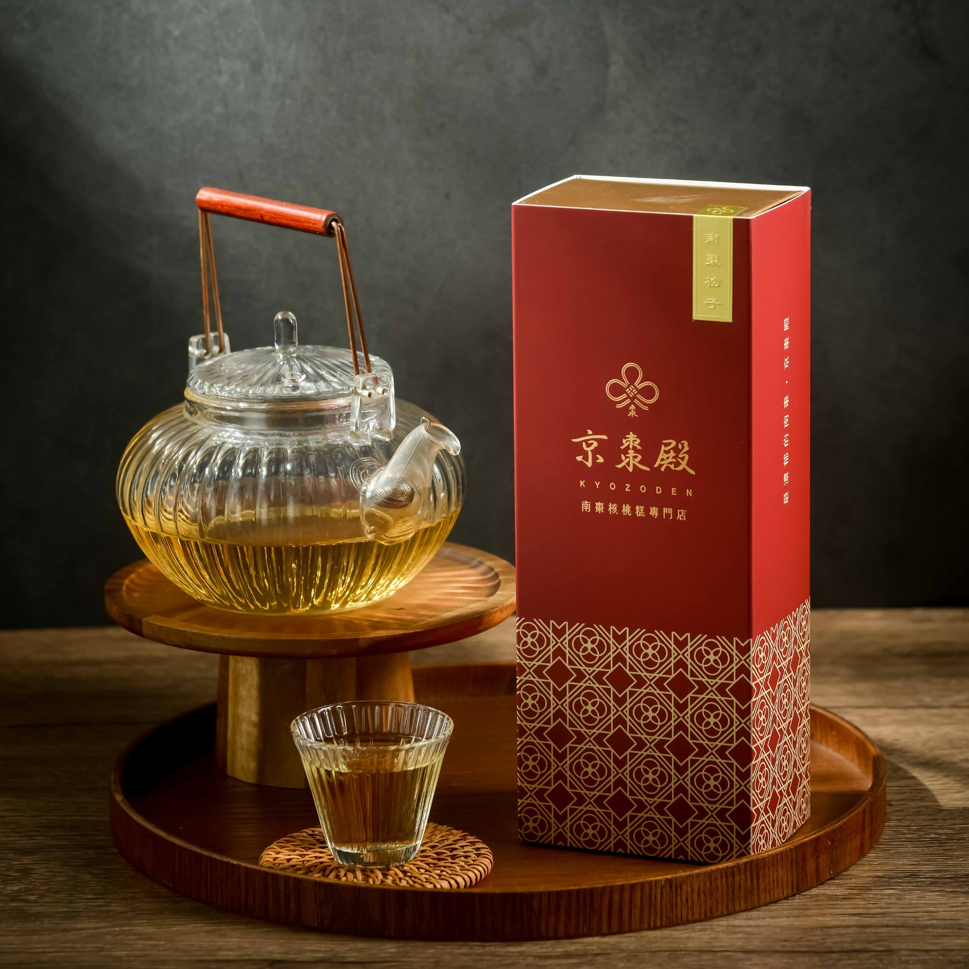 木托盤上放著玻璃茶壺和茶杯，旁邊放著一個紅色禮盒，周圍環繞著茶葉，背景較暗。