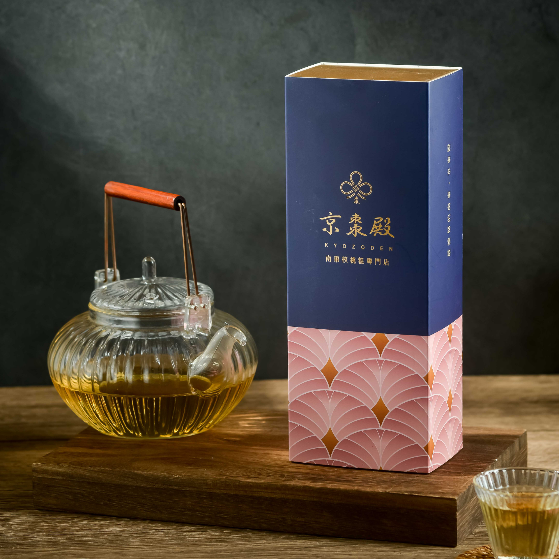 木桌上，玻璃茶壺裡裝著琥珀色茶葉，旁邊放著一個藍粉色的產品包裝盒。