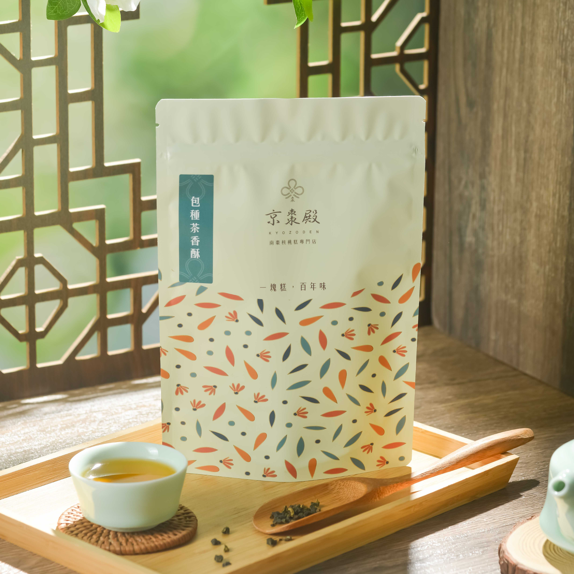 木質表面上放著一個米白色茶包，上面印有色彩繽紛的葉子圖案，旁邊還配有一個茶勺和一小枝薄荷葉。
