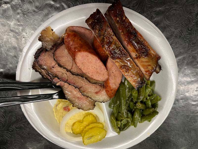 Plate of sliced smoked meat, glazed ribs, green beans, and pickles on a white plate.