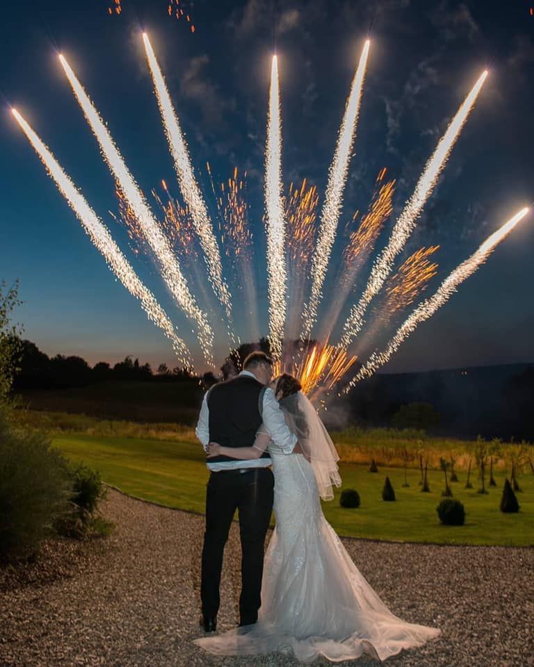 Newlyweds embracing, watching fireworks explode overhead at an outdoor wedding.