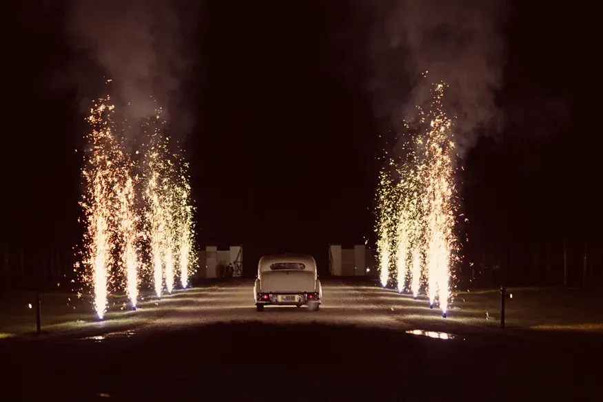 Fireworks erupt along a driveway, framing a car driving toward the viewer at night.