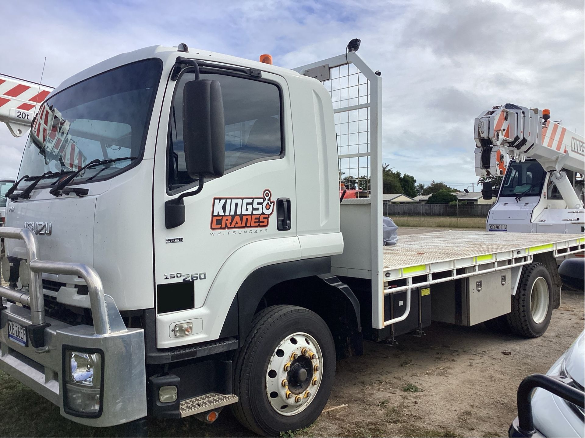 White flatbed tow truck parked on a lot under cloudy skies, with an orange crane in the background - King Cranes in Bowen QLD
