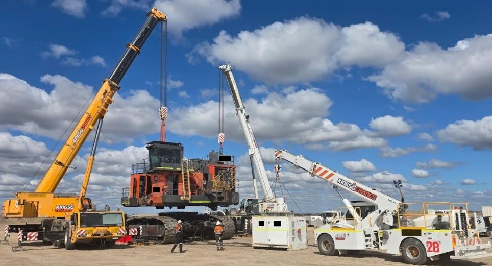 Crane-lifted heavy truck on construction site under blue sky, with workers and equipment nearby