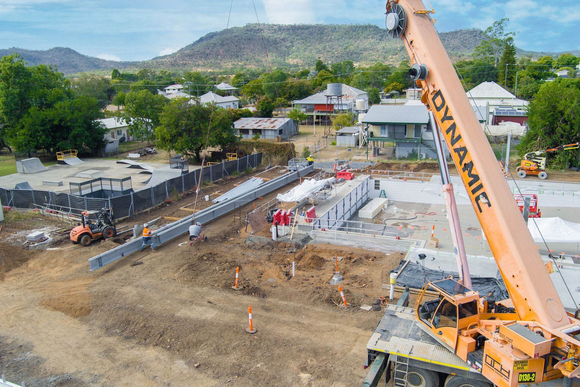 Construction site with crane lifting structure. Orange crane, buildings, trees, and workers — Dynamic Crane Hire In Emerald, QLD