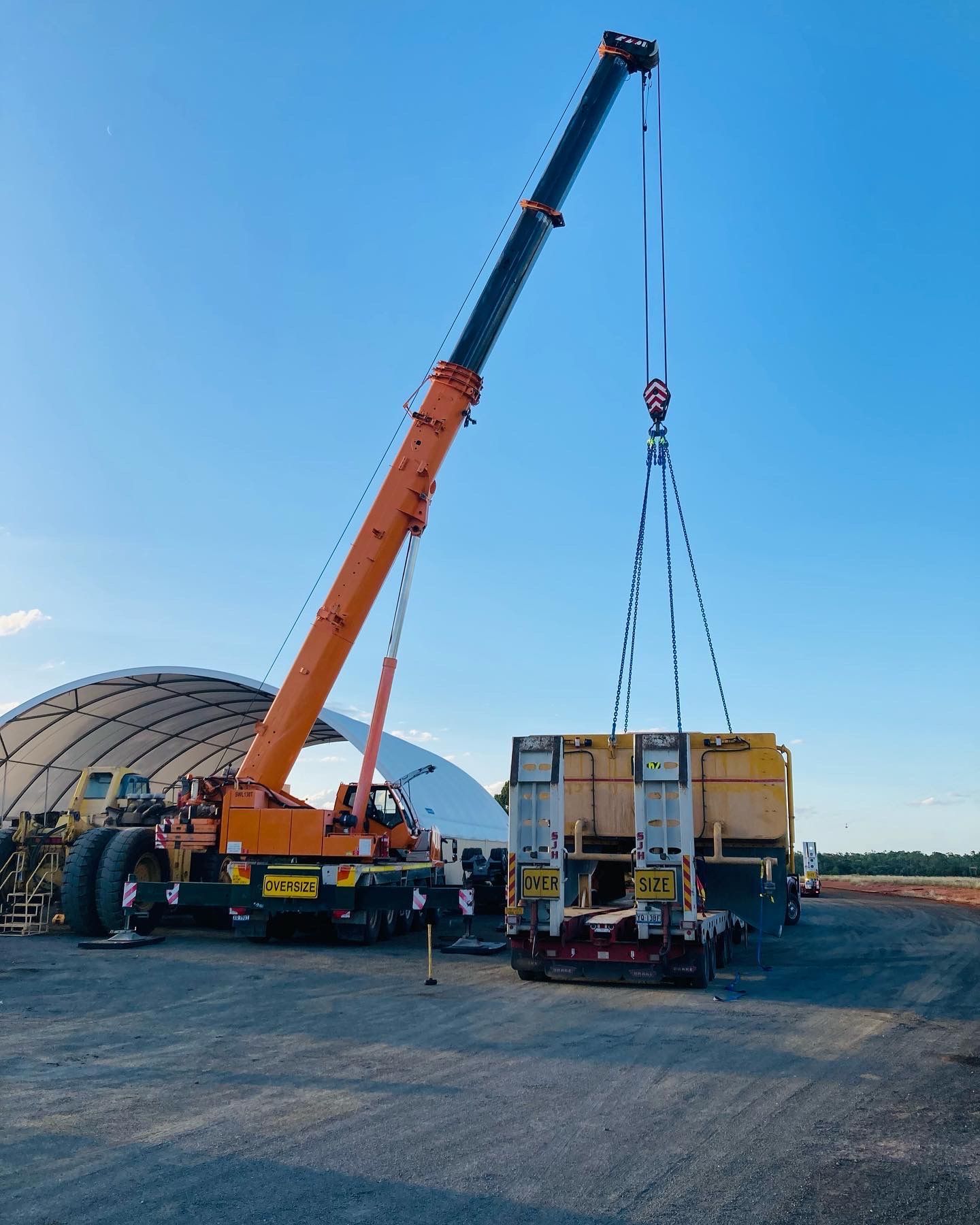 An orange crane lifting a large yellow rectangular object. Blue sky in the background with a white structure  — Dynamic Crane Hire In Emerald, QLD