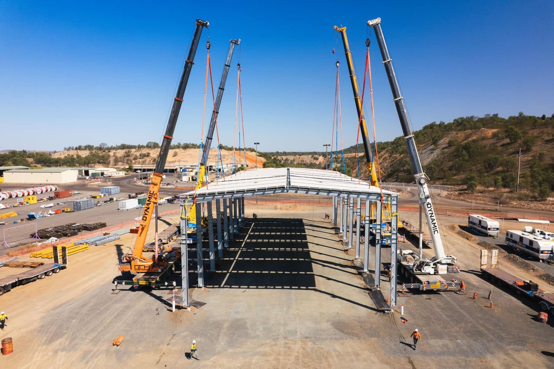 Large cranes lift a curved roof structure onto steel columns at a construction site outdoors on a sunny day  — Dynamic Crane Hire In Emerald, QLD