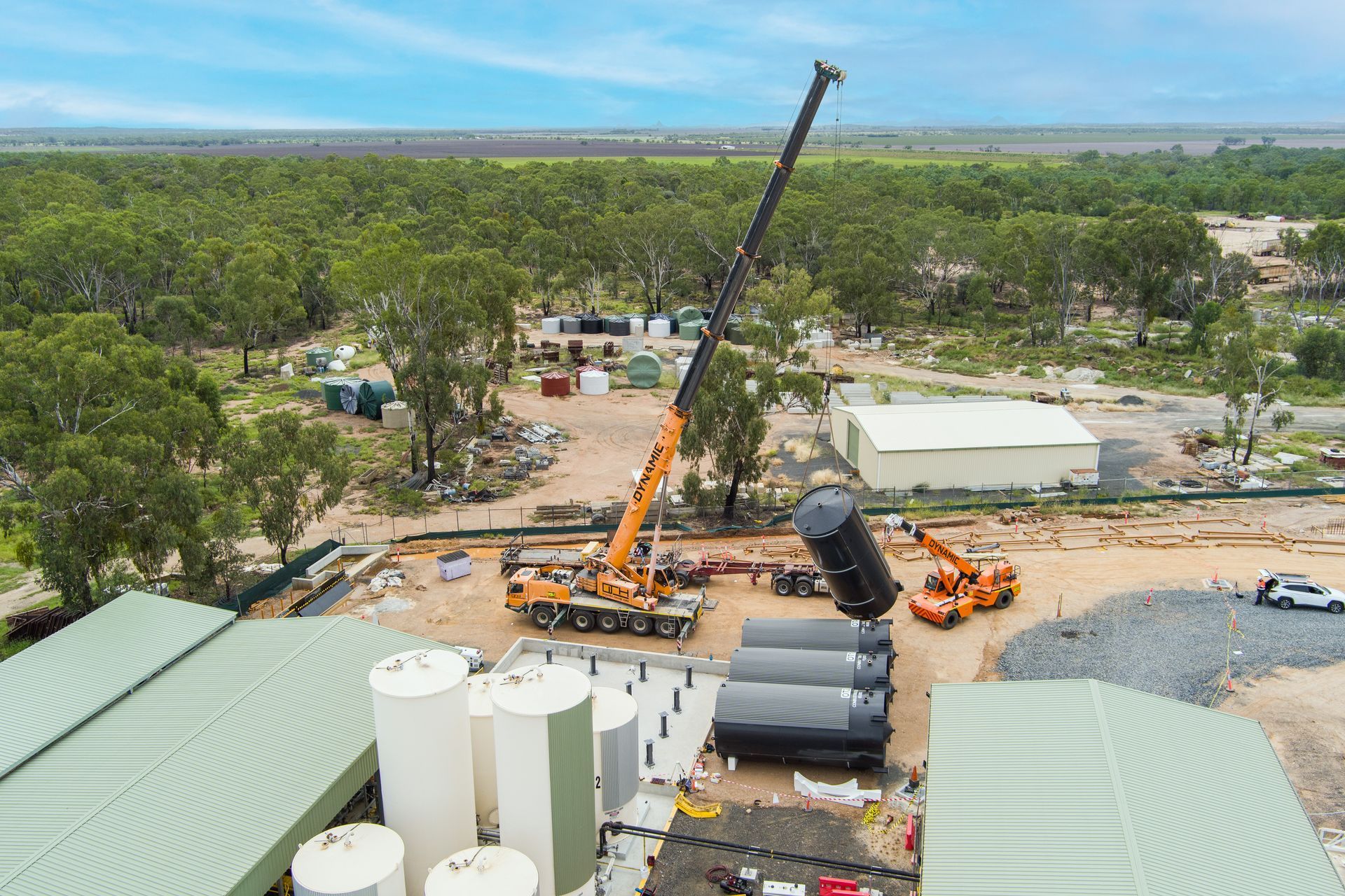 A crane lifting a large black tank at a construction site surrounded by trees and tanks — Dynamic Crane Hire In Emerald, QLD