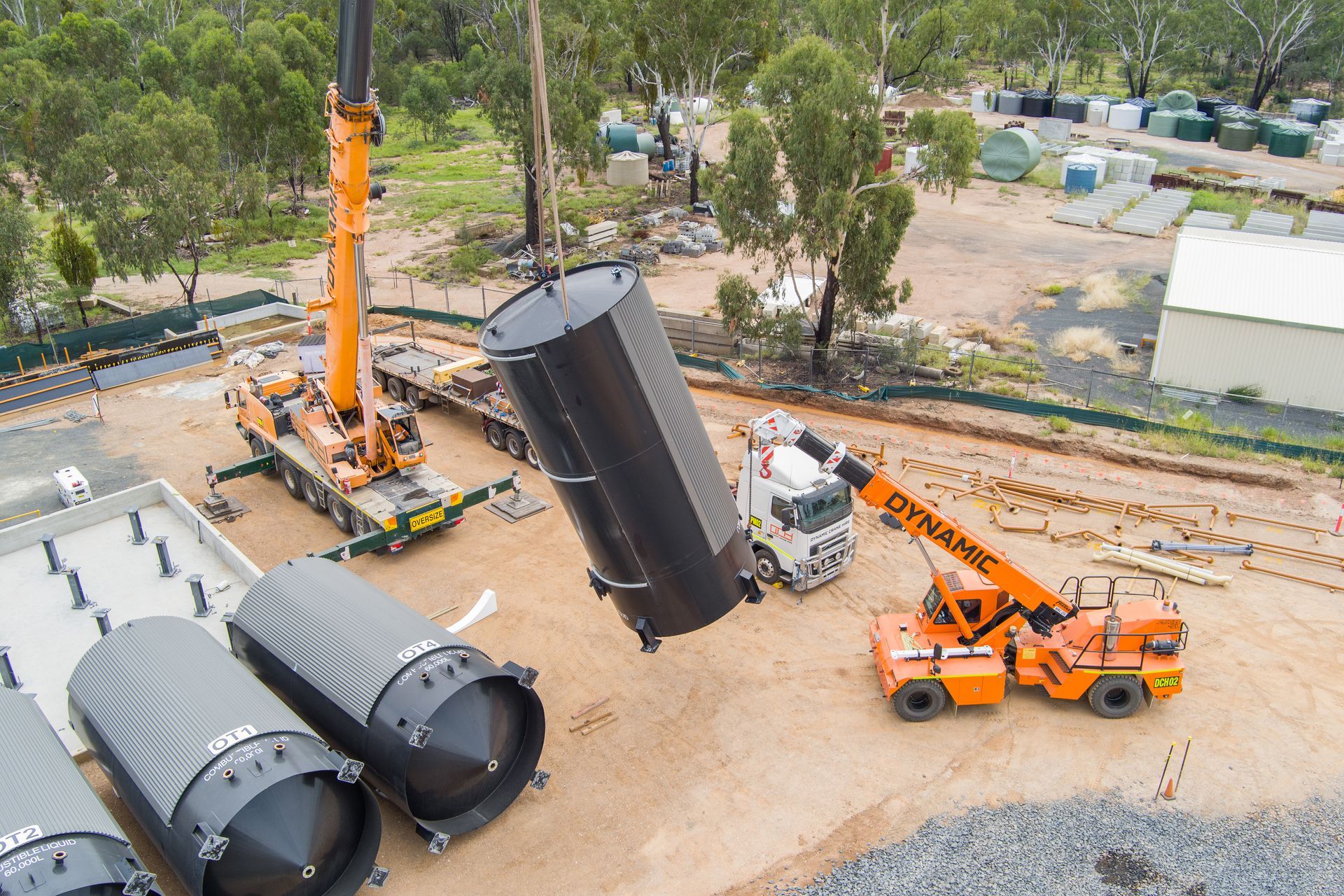 Two cranes lifting a large, black cylindrical tank. Several other similar tanks sit nearby — Dynamic Crane Hire In Emerald, QLD