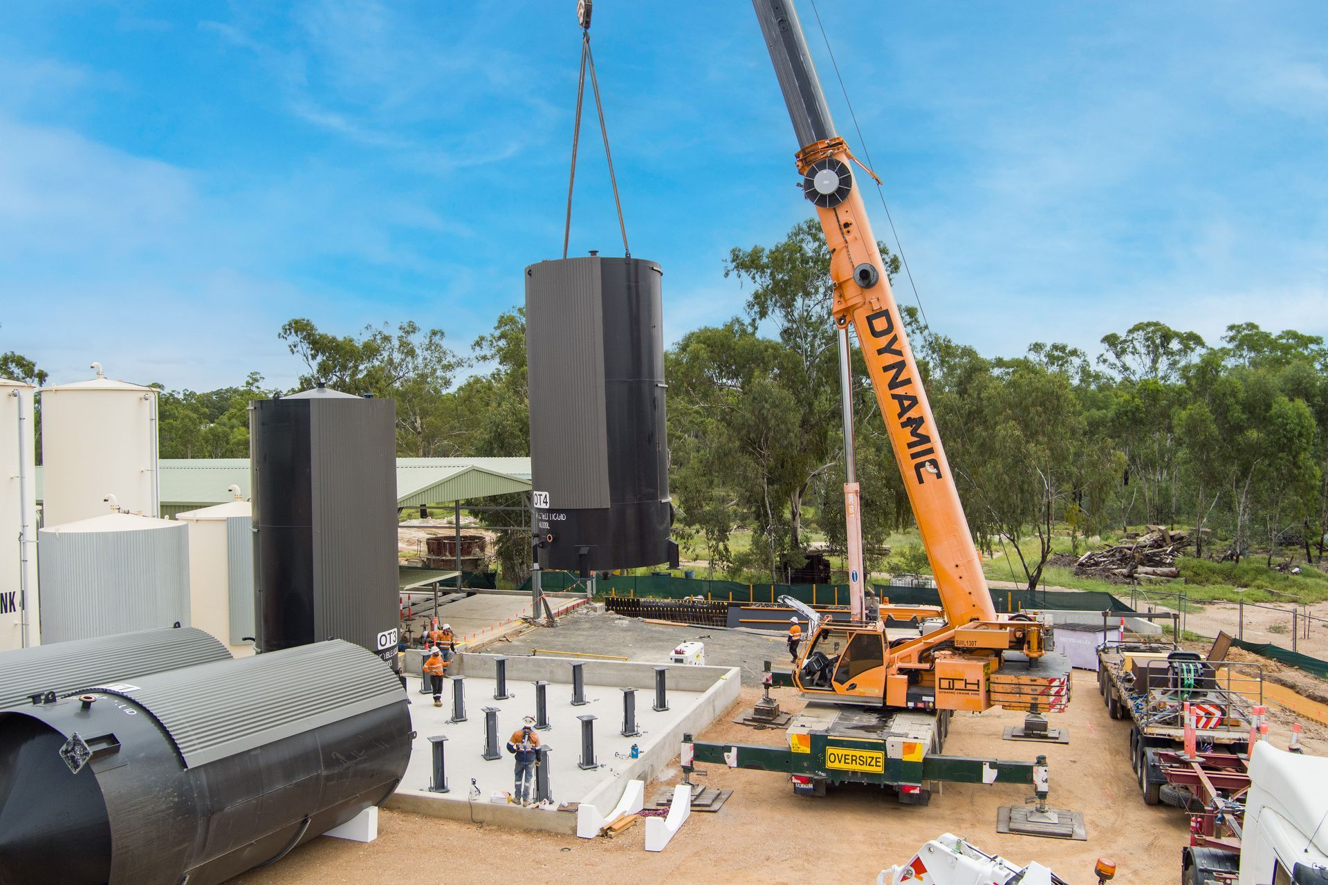 A crane lifts a large black cylinder onto a concrete pad at a construction site. Other structures and people are present — Dynamic Crane Hire In Emerald, QLD