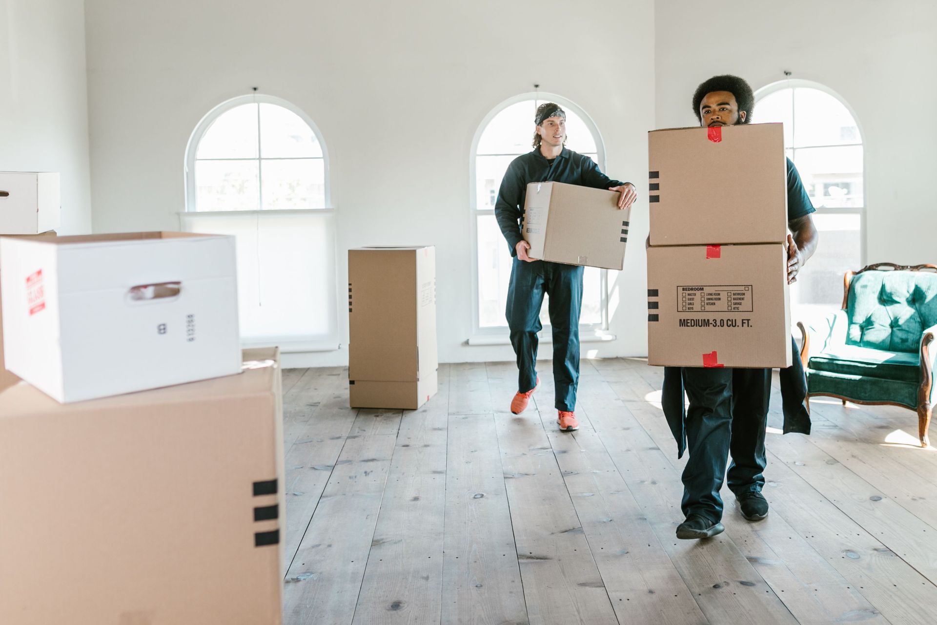 Two people carrying cardboard boxes in a bright, empty room with arched windows and wooden floors