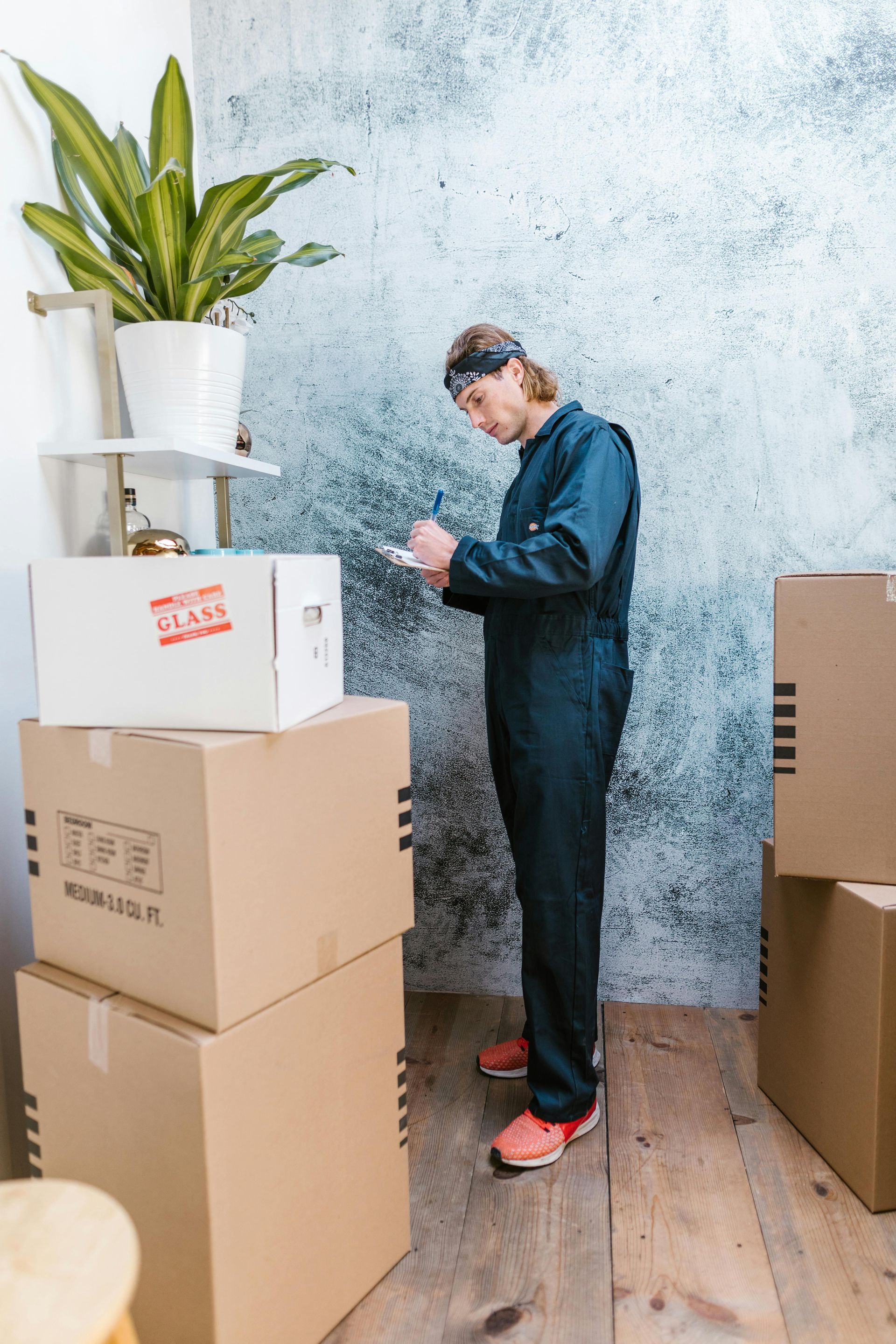 Person in coveralls checking a phone beside stacked moving boxes and a potted plant in a room.