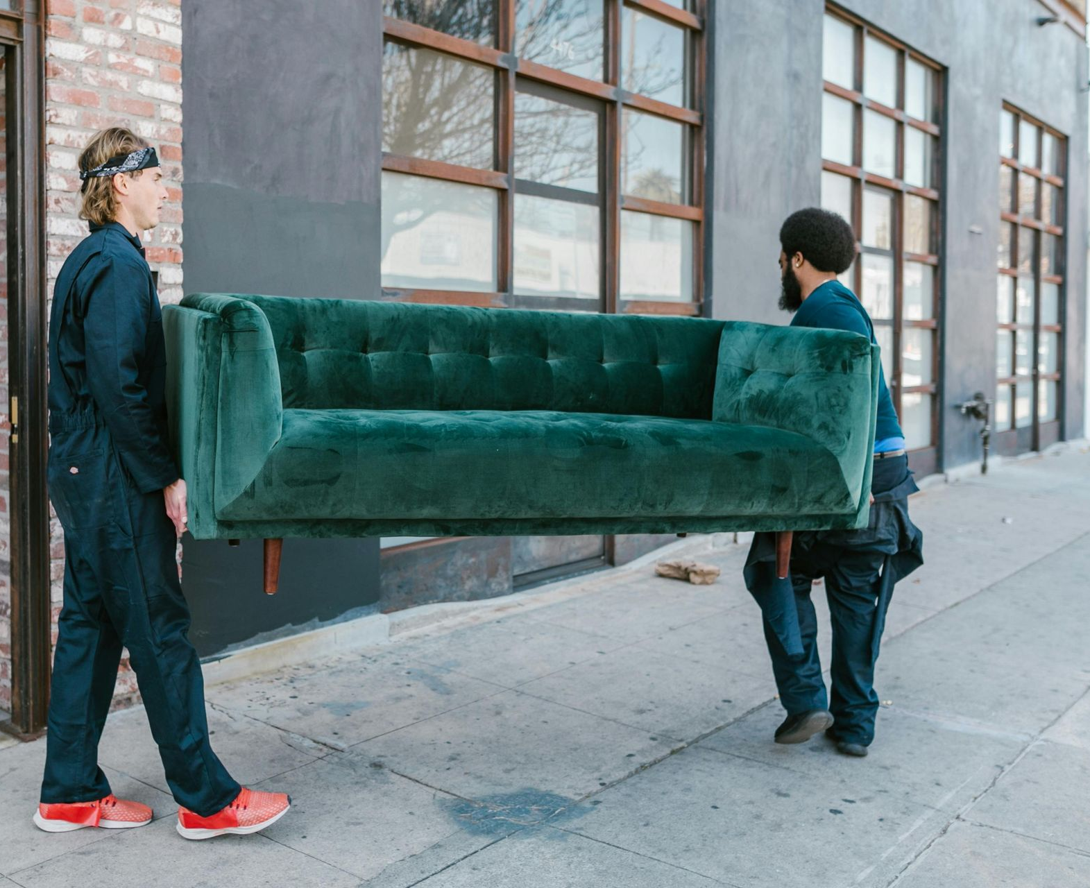 Two people carry a green couch down a city sidewalk beside a building.