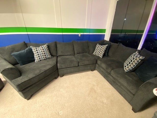 A gray sectional sofa with patterned and solid blue accent pillows sits on a beige carpet against a blue and green wall.
