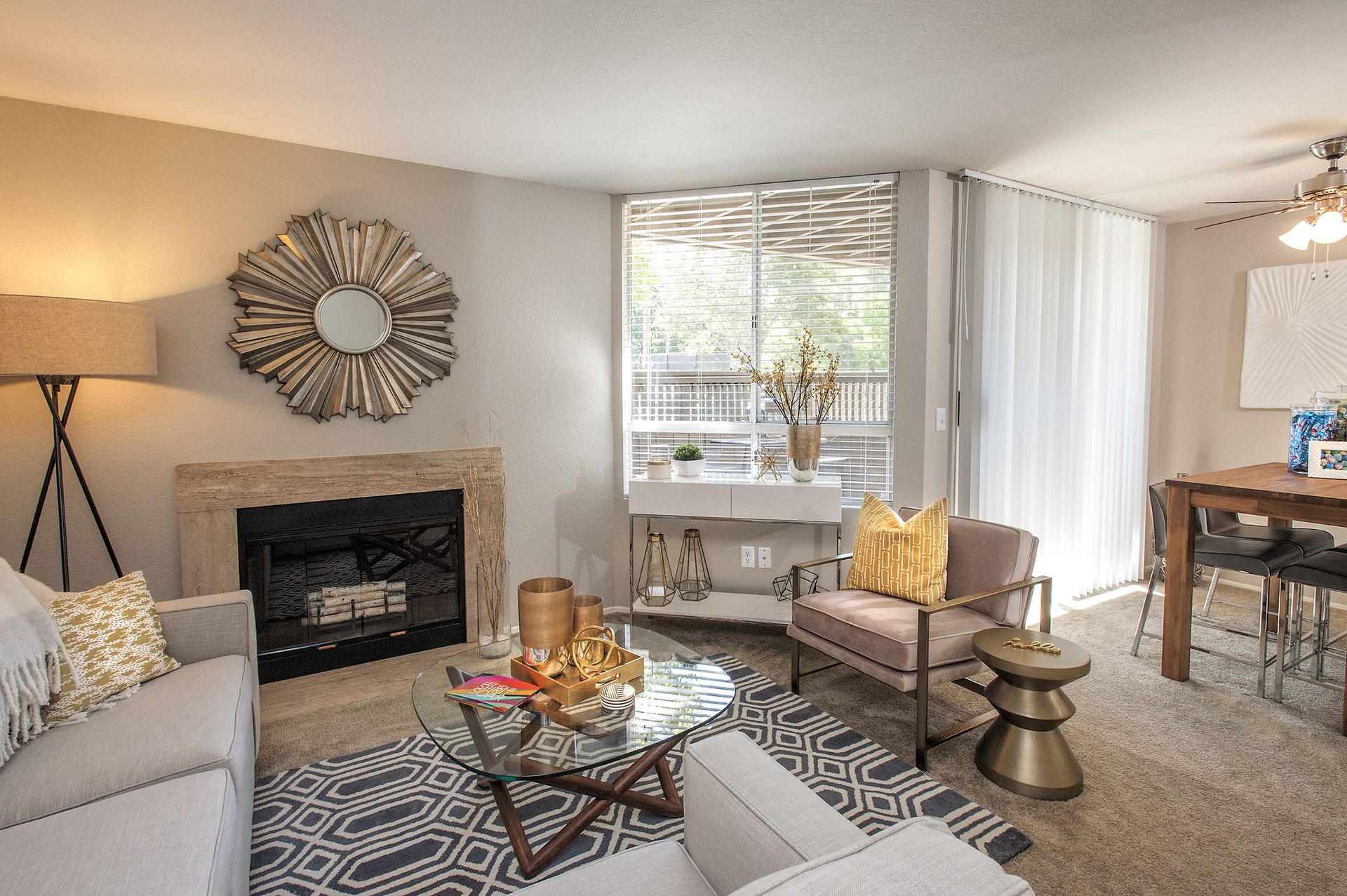 Living room in an apartment with a beige sofa, fireplace, glass coffee table, and a dining area by a large window at Shore Park at Riverlake.