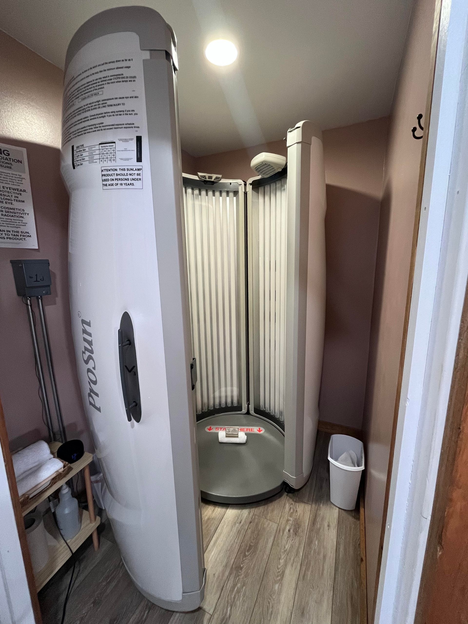 A tanning bed sits in a small room with light pink walls and wood-patterned flooring, alongside a small side table.