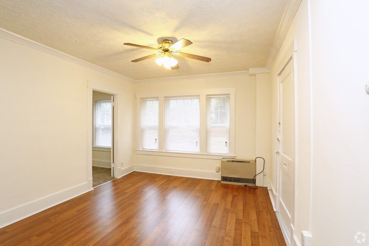 Empty living room with hardwood floors, white walls, ceiling fan, and windows with blinds.