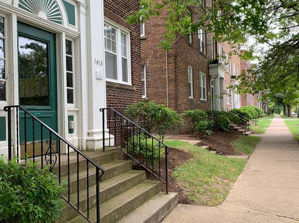 Brick apartment entrance with steps, black railings, and a sidewalk lined by shrubs and trees