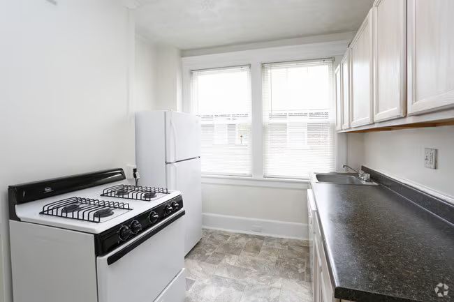 Bright narrow kitchen with white cabinets, black countertop, gas stove, and a window at the end.