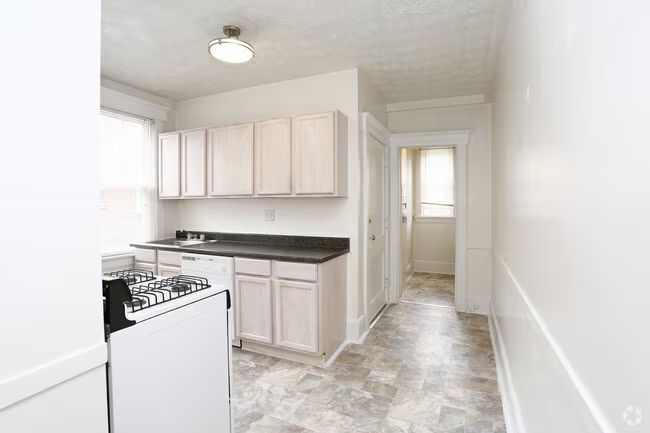 Bright narrow kitchen with white cabinets, gas stove, black counters, and a hallway beyond.