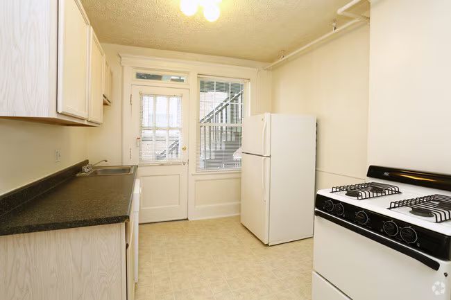 Small beige kitchen with white cabinets, black countertops, stove, refrigerator, and a door to an exterior stairway.