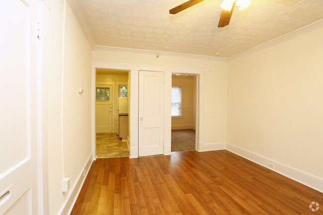Empty room with hardwood floors, white walls, and a ceiling fan, viewed toward a hallway and doorway.