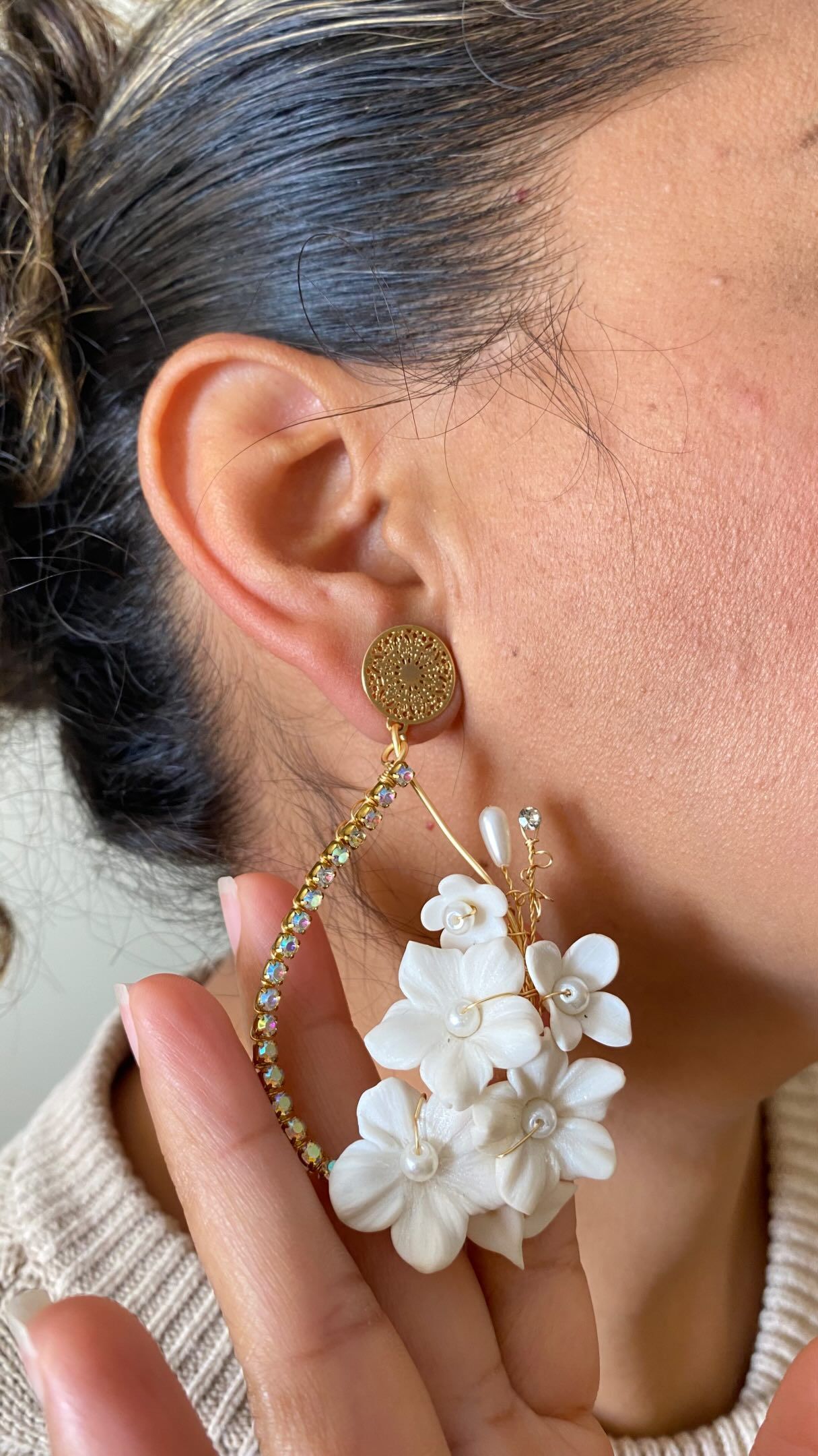 Primer plano de una mano sosteniendo un pendiente colgante floral dorado y blanco junto a una oreja