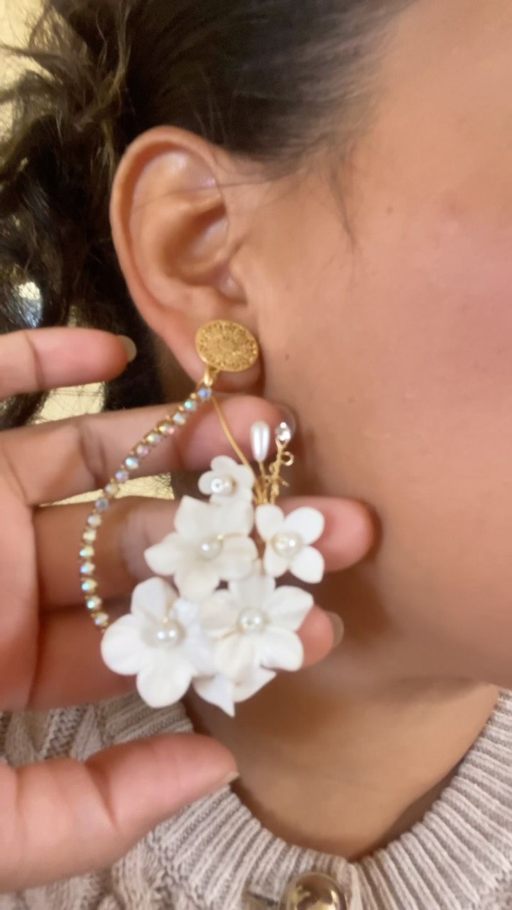 Una mano sostiene un pendiente blanco con forma de flor cerca de la oreja de una persona, mostrando un cierre dorado y detalles de perlas