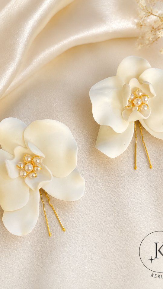 Tela de satén color crema con dos horquillas para el pelo en forma de flor blanca, con detalles dorados en los tallos y los centros