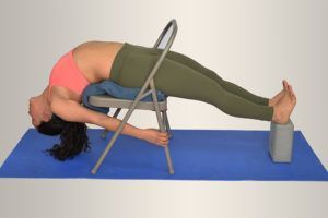 Mujer en pose de yoga usando una silla y bloques, espalda arqueada, brazos extendidos hacia abajo, vistiendo leggings y una camiseta rosa, sobre una colchoneta azul.