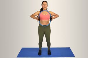 Mujer usando el anillo de Pilates, de pie sobre una colchoneta, con las manos en las manijas del anillo, contrayendo los músculos del pecho.