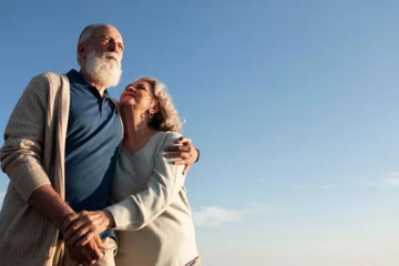 Una pareja de ancianos, un hombre con barba blanca y una mujer con el brazo alrededor de él, mirando hacia el cielo azul brillante.