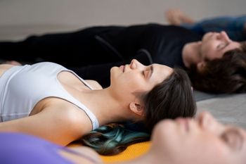 Personas tumbadas sobre colchonetas de yoga, con los ojos cerrados, con aspecto relajado. Interior.