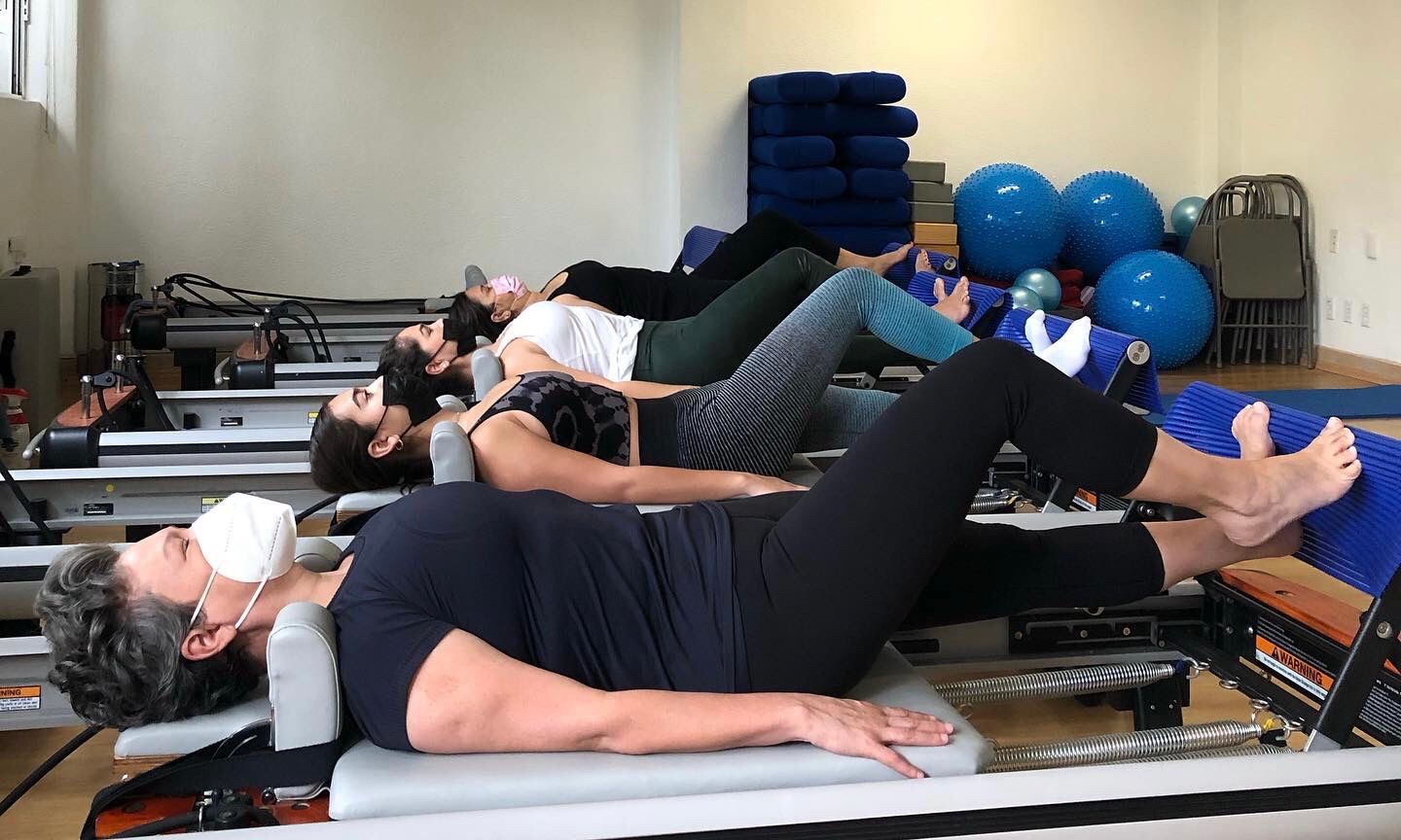 Personas en máquinas de pilates en un estudio, haciendo ejercicios de piernas. Una persona lleva mascarilla.