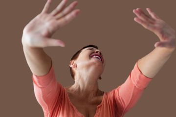 Mujer de pelo corto riendo, con los brazos alzados. Camisa rosa y fondo marrón.