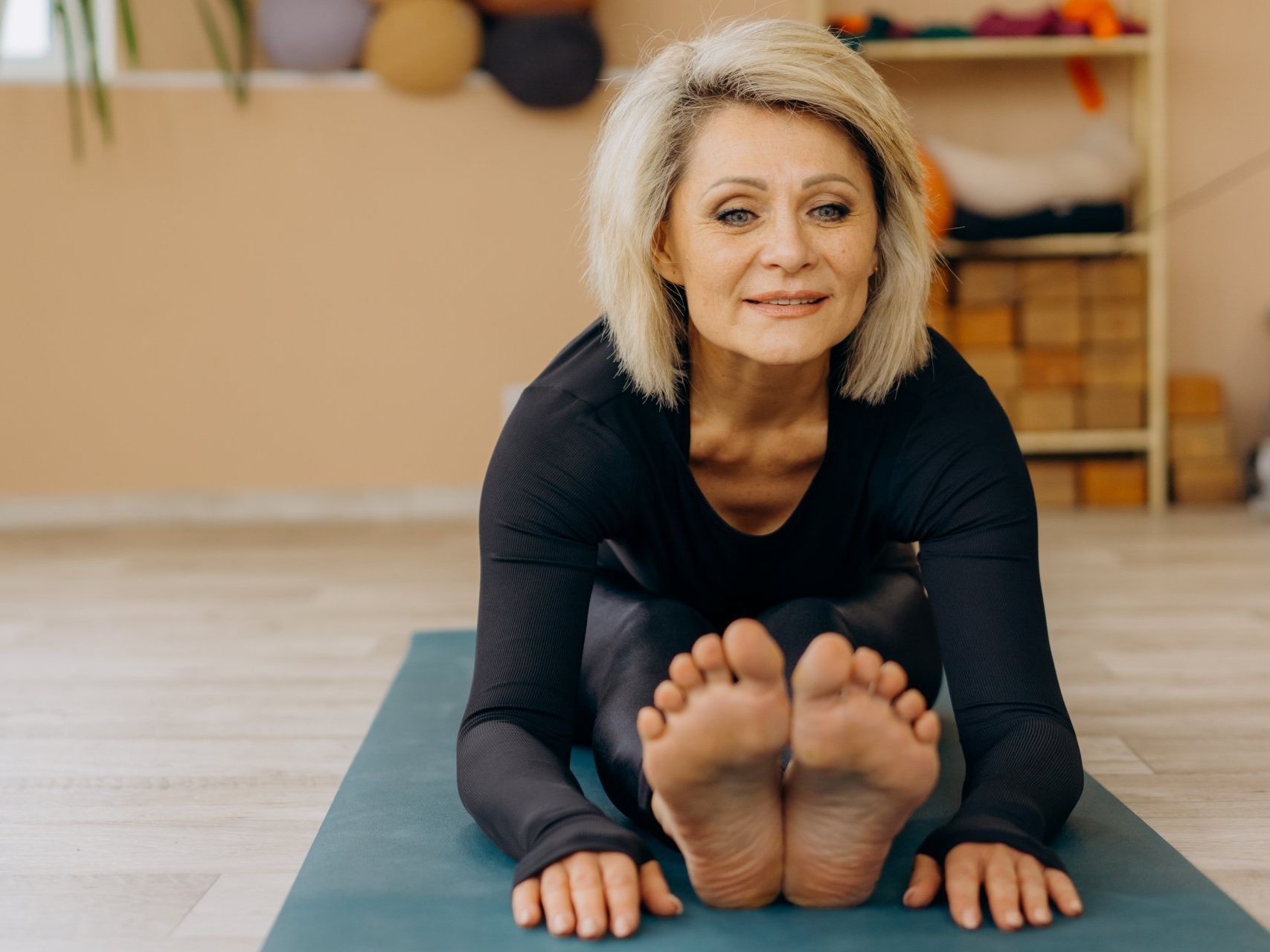 Mujer estirando su cuerpo sobre una esterilla de yoga, en interiores. Sonríe y viste ropa deportiva negra.