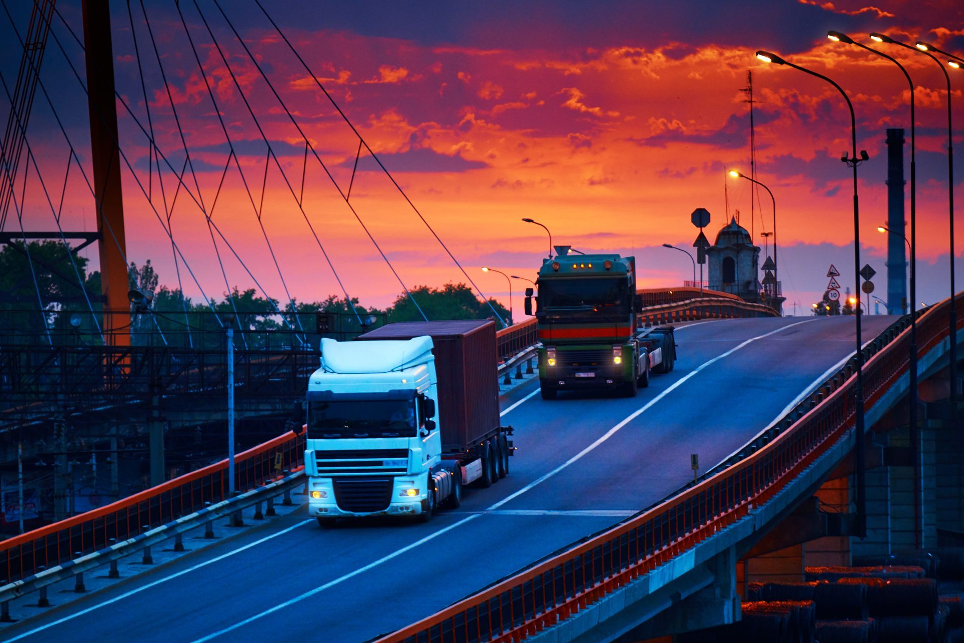 Two trucks driving on a bridge at sunset under a vivid purple-orange sky.