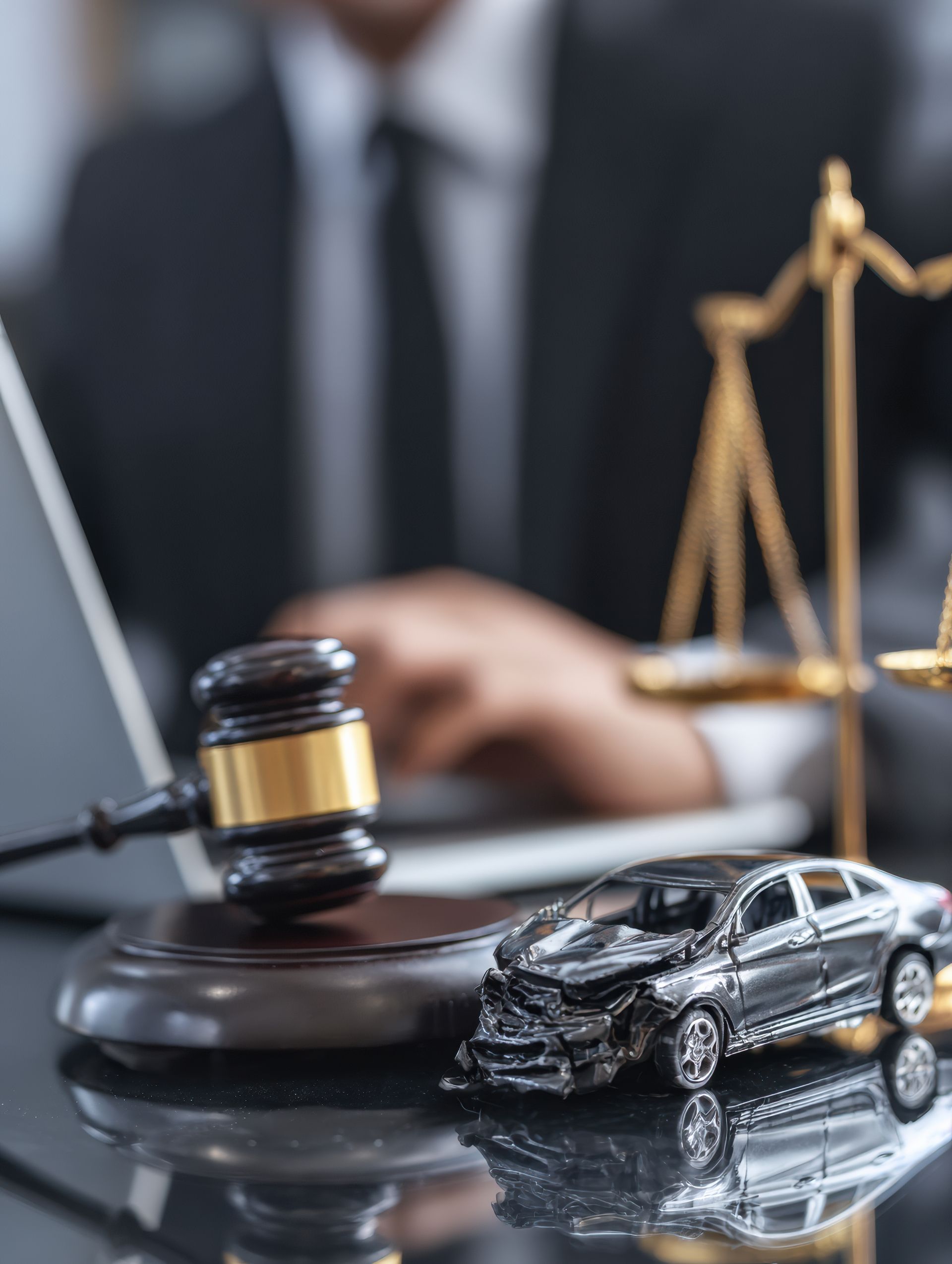 Miniature damaged car next to gavel and scales; a person in a suit is working on a laptop.