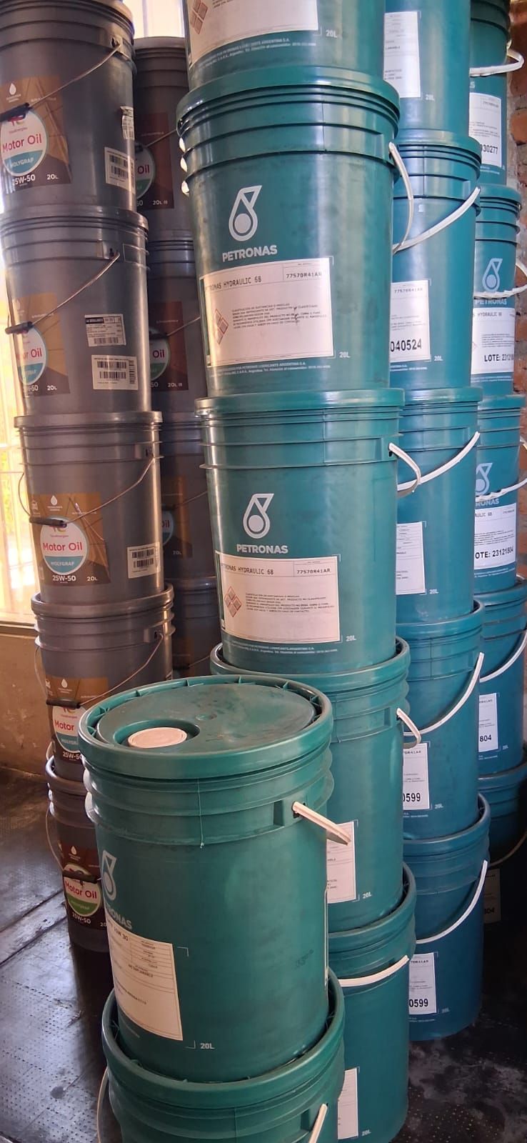 Pilas de bidones metálicos de color verde azulado y marrón, apilados en posición vertical en un pasillo de un almacén.
