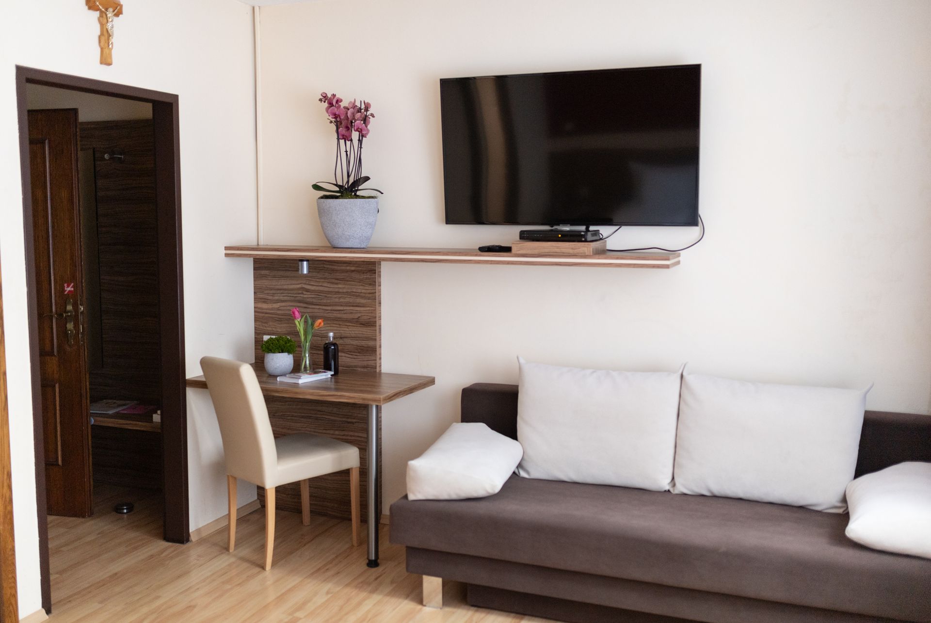 Zimmer in Hotel Maria, Brauner Holzboden, teils braune Wandverkleidung. Im Fokus steht der kleine Wandtisch und ein Sessel. Daneben eine Couch und darüber ein Fernseher. 