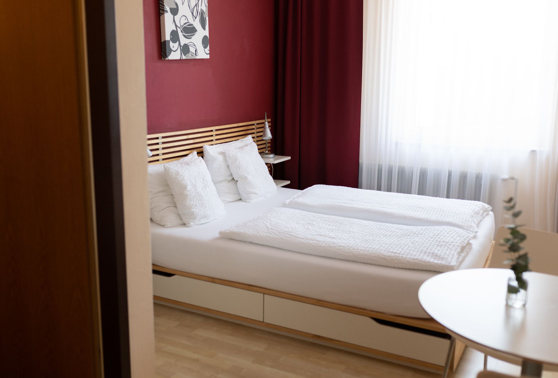 Zimmer Hotel Maria, 1,40m Doppelbett mit weißen Laken. Kopfteil steht an einer teils Holzverkleideten, teils rot gestrichenen Wand.