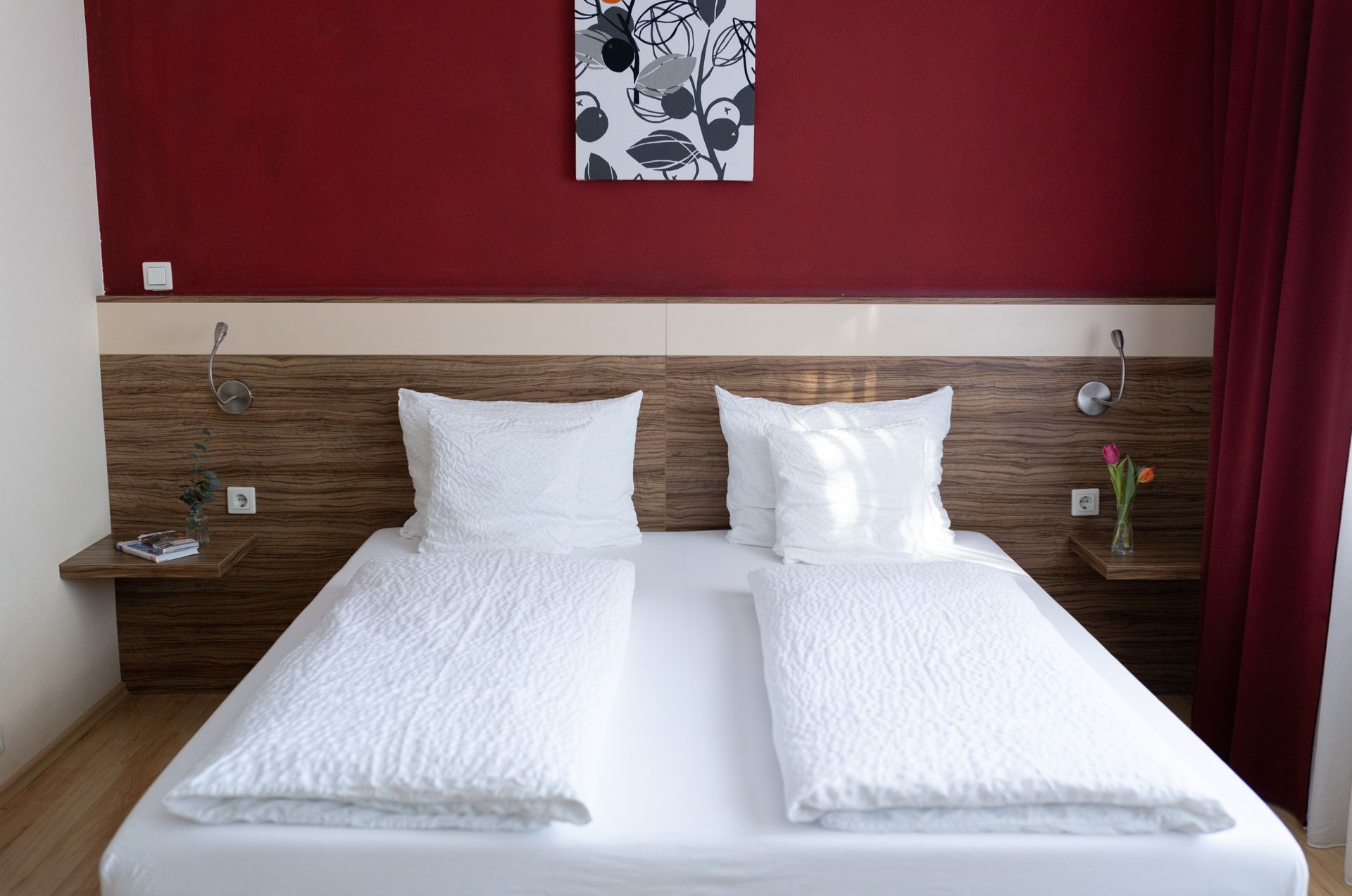 Zimmer Hotel Maria, Doppelbett mit weißen Laken. Kopfteil steht an einer teils Holzverkleideten, teils rot gestrichenen Wand.