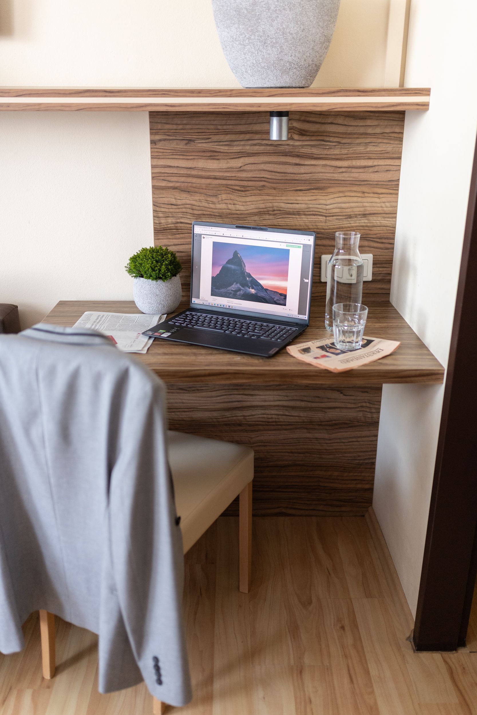 Zimmer in Hotel Maria, Brauner Holzboden, teils braune Wandverkleidung. Im Fokus steht der kleine Wandtisch und ein Sessel. Auf dem Tisch steht ein offener Laptop.